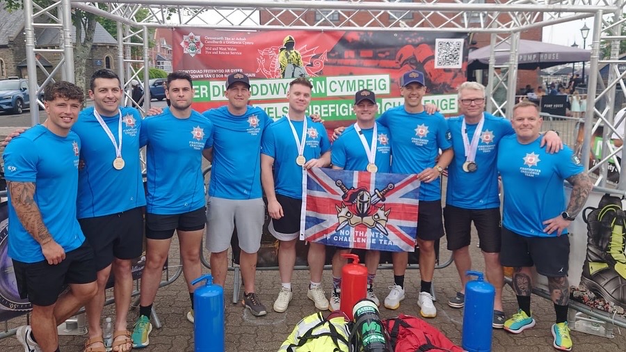 The Northamptonshire Fire and Rescue Service team at the Welsh Firefighter Challenge are shown - all nine of them wearing blue shirts with the Fire Service logo on. Three of them in the middle are holding a flag with the words Northants FF Challenge Team, written over a Union Jack.
