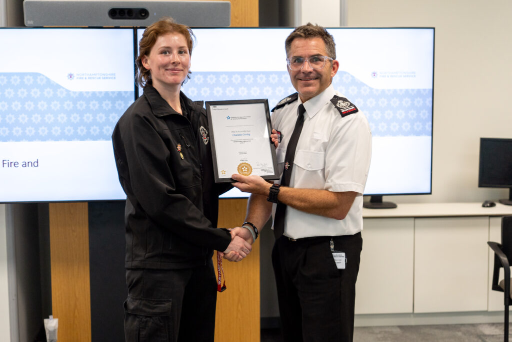 Pictured on the left, wearing a black shirt and trousers, is firefighter Charlotte Cowley. She is being presented with her Apprenticeship certificate by Deputy Chief Fire Officer Simon Tuhill. who is shown on the right in a white shirt. They are pictured in front of a digital display showing the Northamptonshire Fire & Rescue Service branding.