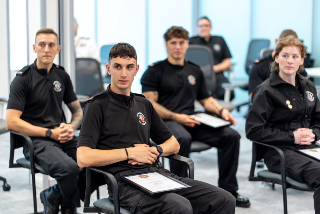 A number of firefighters are shown sitting down wearing black shirts. They are the first cohort of firefighters to gain apprenticeship qualifications through Northamptonshire Fire & Rescue Service.