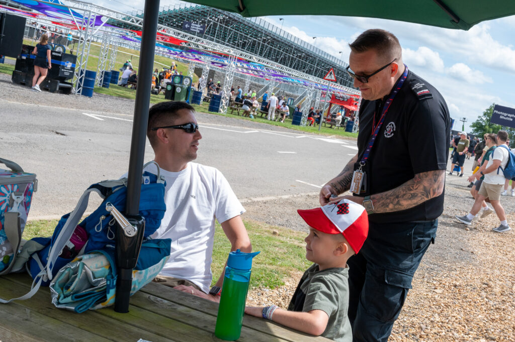 A Northamptonshire firefighter in black, picture on right wearing sunglasses, is shown speaking to a man in a white shirt and sunglasses. In the foreground is a young boy wearing a red and white cap. The firefighter is giving safety advice to the man.