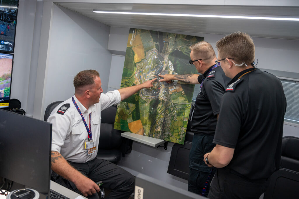 Group Commander Matt Butler, shown on the left in white, points at a map of the Silverstone circuit while in discussion with two firefighters wearing black, one of whom is also pointing at the map of the race track.