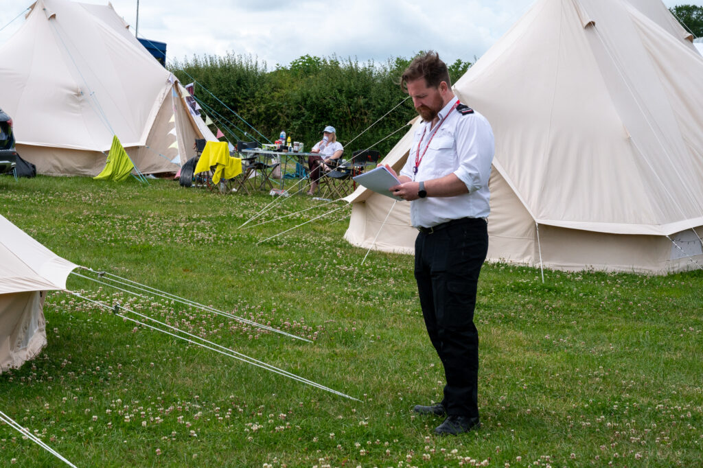 A Fire Protection Officer wearing a white shirt is shown looking at his notes following an inspection of a campsite. There are two white tents in the background.