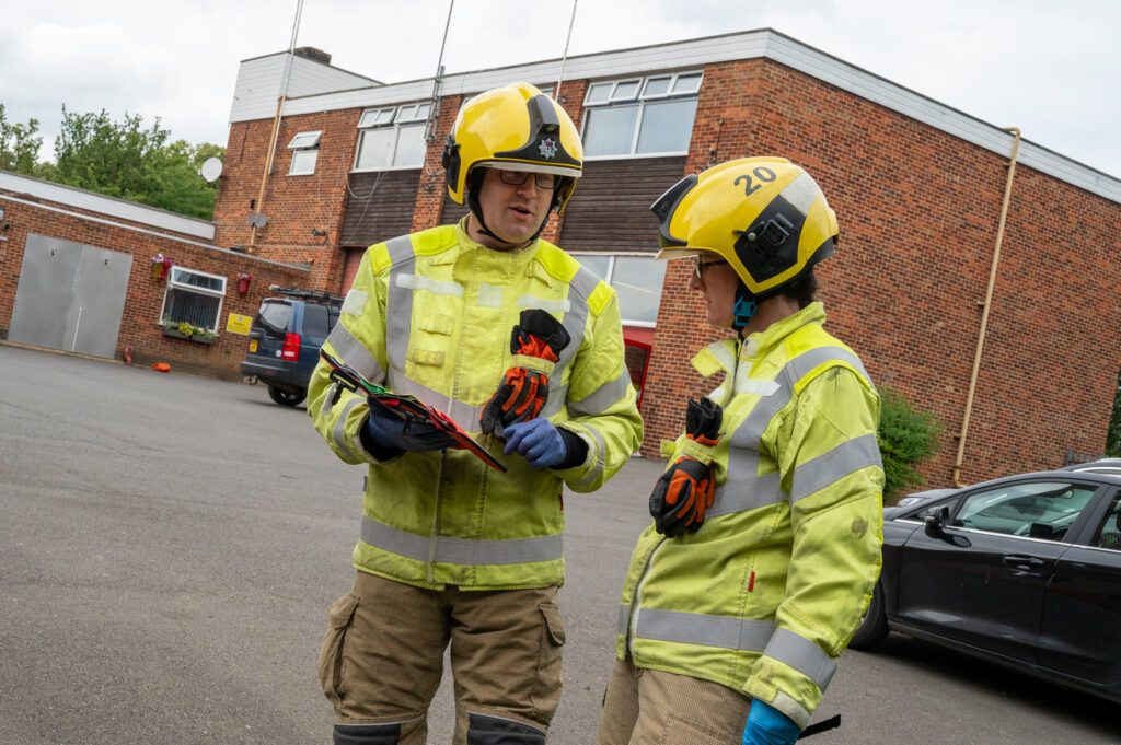 Two firefighters wearing high visibility jackets and yellow helmets are shown discussing how to triage a patient