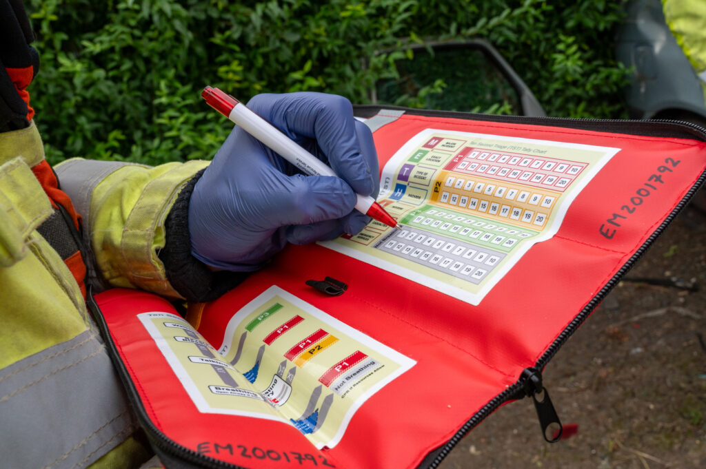 A firefighter in a high-visibility jacket writes with a pen on the red triage kit