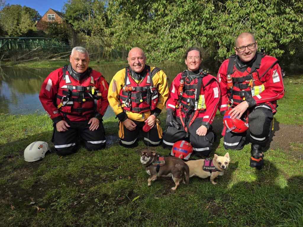 Four firefighter in water safety equipment kneeling down on the grass near the waters edge with two dogs they rescued