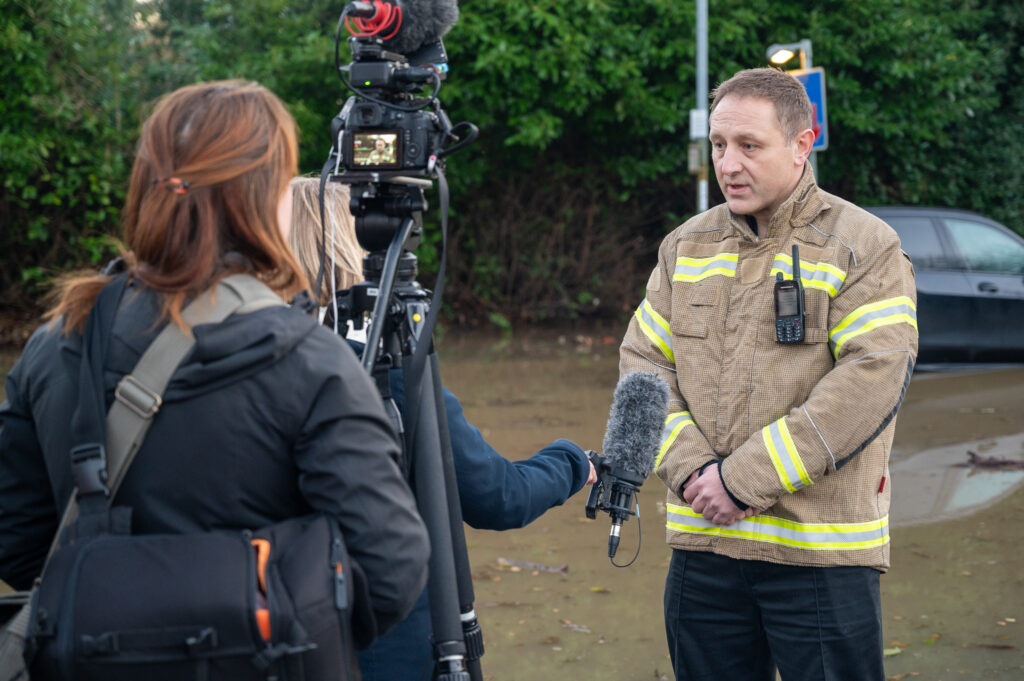 Area Commander Neil Sadler speaks to a news reporter in front of a camera, next to flood water and a stranded car