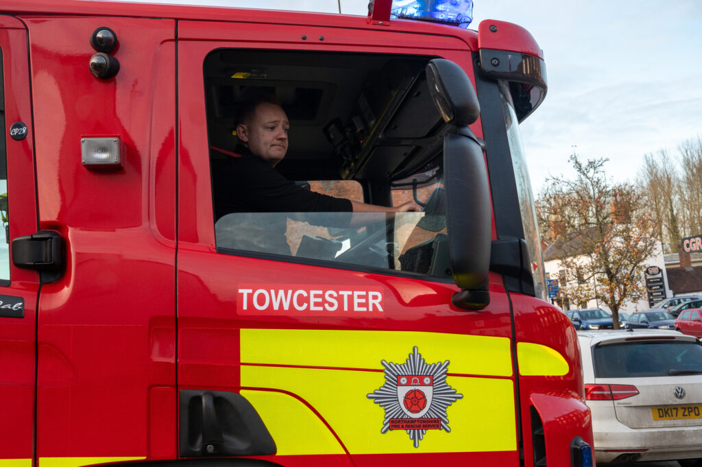 A firefighter in black kit driving a red fire engine with TOWCESTER on the side