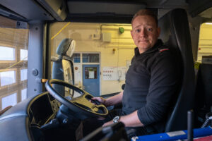 A firefighter in black uniform behind the wheel of a fire engine