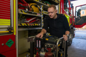 A firefighter in black looking at some kit on the side on a fire engine