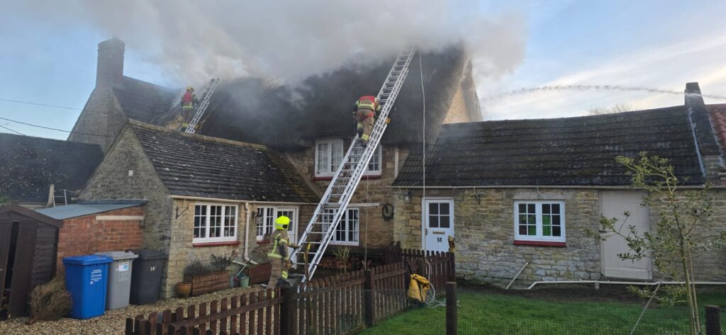 Three firefighters up ladders tackle a fire in a thatched roof property