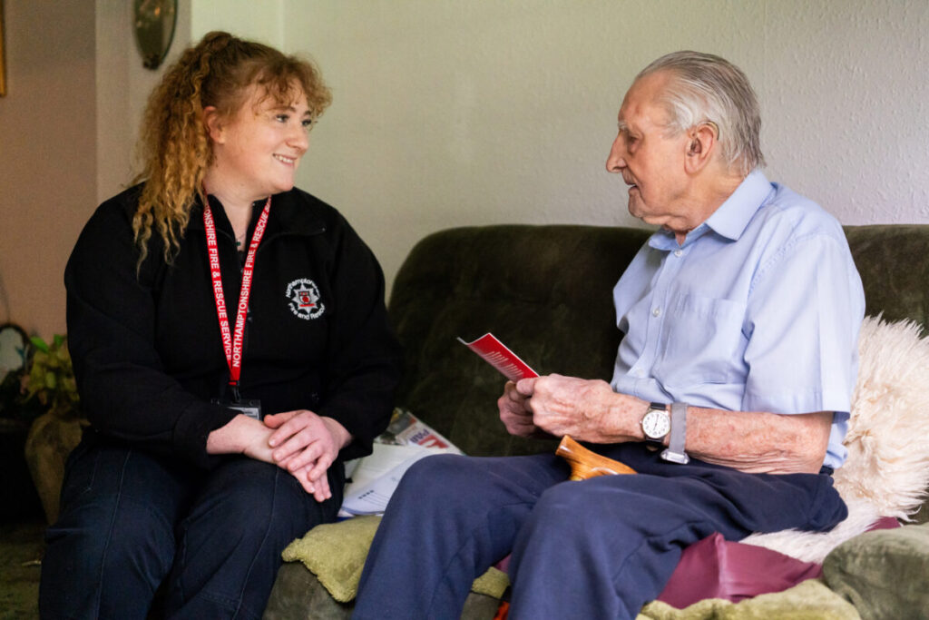 2 people sitting on a osfa discussing fire safety in the home, one is wearing NFRS uniform and lanyard